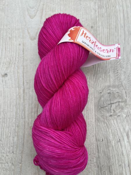 Handgefärbter Wollstrang in kräftigem Pink (Herzfasern 2.0) auf heller Holzoberfläche. Das Garn ist gleichmäßig gefärbt und zeigt eine glatte, feine Struktur. Banderole mit dem Schriftzug „Herzfasern – Garnkunst & Strickliebe“.